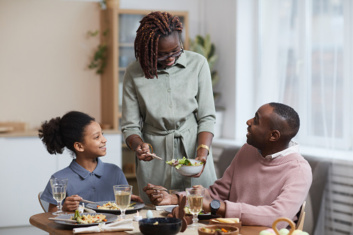 Portrait of modern African-American woman serving food for family while enjoying dinner together in cozy home interior, copy space