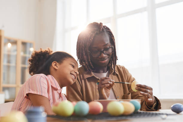 madre e figlia afroamericane che dipingeno uova di pasqua alla luce del sole - child candid indoors lifestyles foto e immagini stock