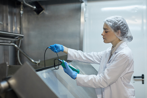 Side view portrait of young female worker holding temperature meter while probing mixture at industrial food factory, copy space