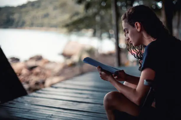 Young woman reading a book sitting on a wood balcony by the beach at dusk