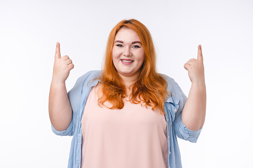 Adorable young plus size woman pointing at copy space isolated in white