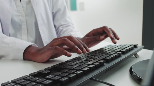 Close up of hands of young African woman in medical uniform typing on keyboard in hospital. Good-looking female nurse sitting at table and working on computer. Technology, health, medicine concept