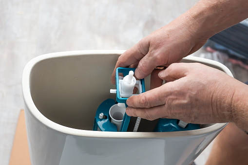A plumber installs a water pump in a ceramic toilet cistern. Drainage system installation, home repair.