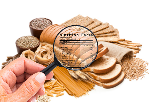 Close up view of a female hand holding magnifying glass on a group of carbohydrates food to look for the details of the nutrition facts of this type of food. A Nutrition Facts label is visible on the magnifying glass.