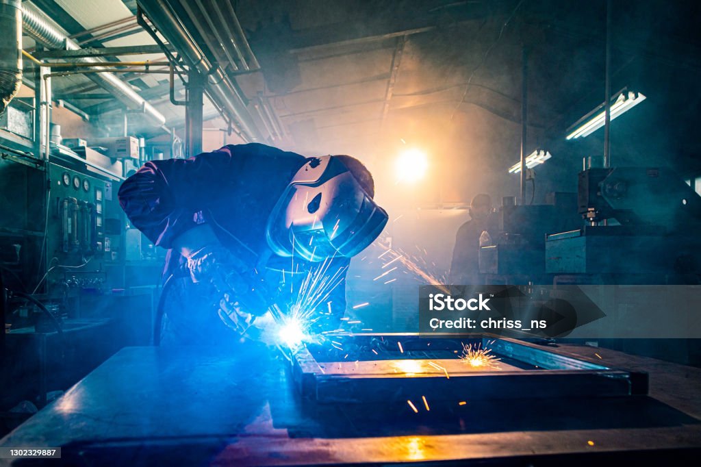 Two handymen welding and grinding metal at workshop The two handymen performing welding and grinding at their workplace in the workshop, while the sparks "fly" all around them, they wear a protective helmet and equipment. Welder Stock Photo