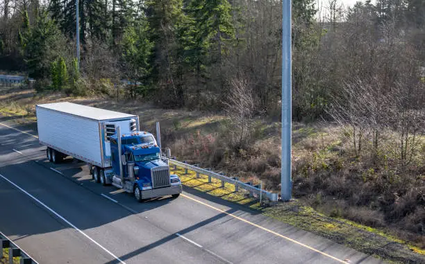 Photo of Classic blue big rig semi truck tractor with chrome accessories transporting frozen cargo in refrigerator semi trailer running on the left line of multiline highway with one way traffic direction
