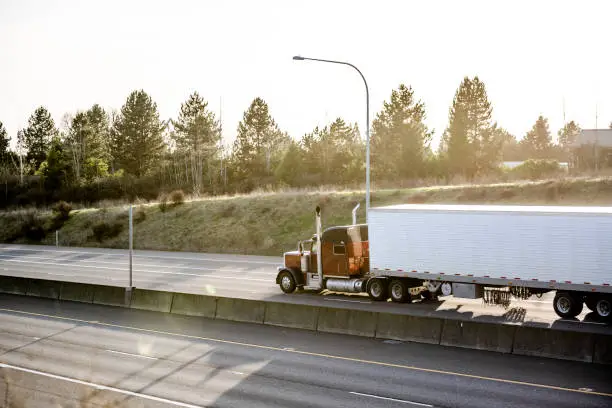 Photo of Brown big rig classic semi truck with refrigerator semi trailer running with cargo on divided highway road in sun light