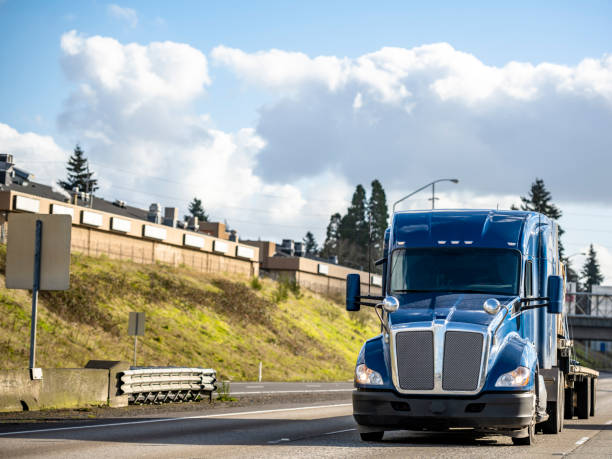 istock Powerful blue big rig bonnet semi truck tractor transporting cargo on step down semi trailer running on the wide highway with hill and traffic road signs 1302331167