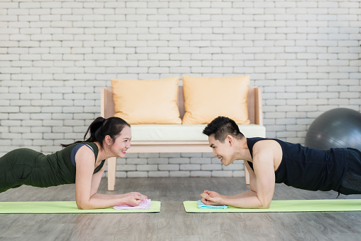 Happy asian couple doing plank exercise together in living room at home. Sporty woman and man smile and make exercise on yoga mat indoors daily routine while social distancing. Home fitness concept.