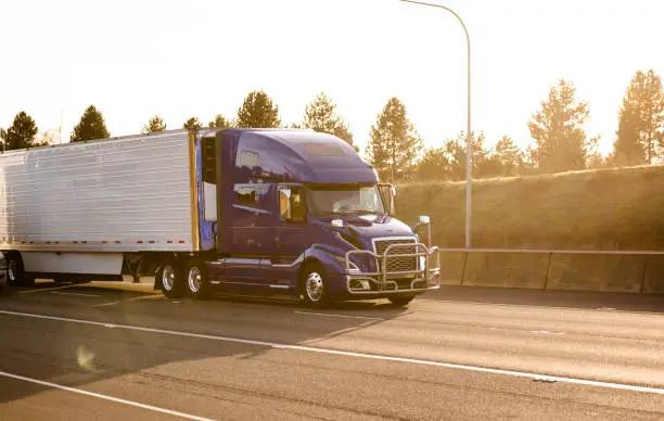 Photo of Dark blue big rig semi truck with grille guard transporting refrigerated semi trailer with frozen cargo running on the sunny divided highway road