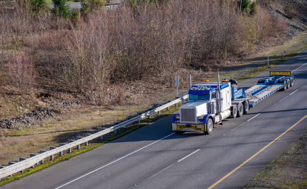istock White and blue classic big rig semi truck with empty step down semi trailer and oversize load signs on the front and back moving on the one way highway road with bare winter trees on the hill 1302332388