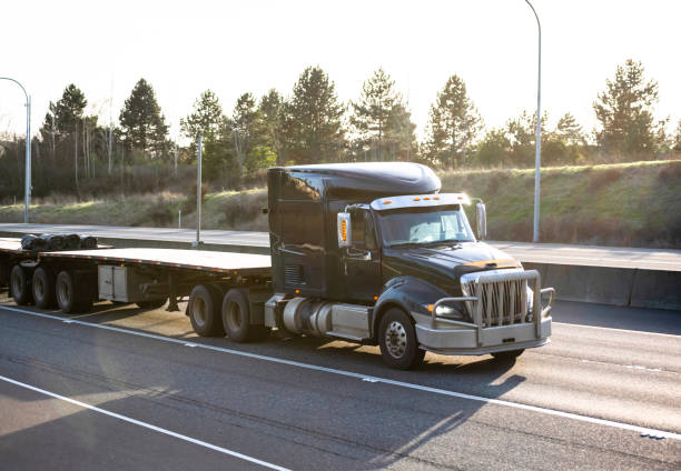 istock Black big rig low cabin semi truck with two empty flat bed semi trailers running on the highway road at sunlight 1302333240