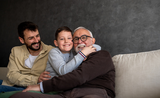 Grandfather is hugging his grandson and grandson is hugging him back. By him sits his father enjoying in time he spends with his family.
