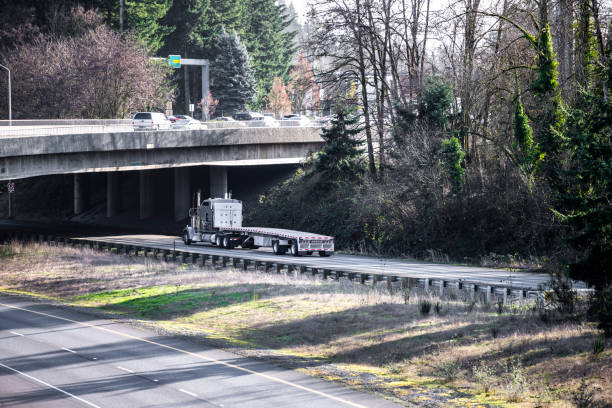 istock Gray big rig classic semi truck with empty flat bed semi trailer running under the bridge across the divided highway road intersection 1302333586