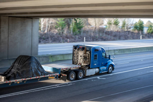 istock Powerful big rig blue semi truck tractor transporting covered cargo on step down semi trailer running on the wide highway road with bridge 1302334122