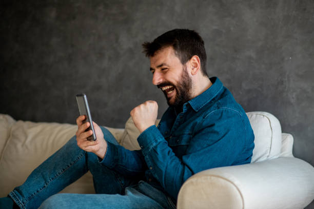 istock Bearded young man laughing and reading text message. He is sitting on sofa in living room. 1302334419