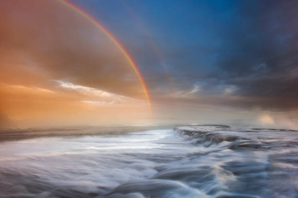 istock Ocean waves flowing over rocks at sunrise with rainbow and storm 1302339847