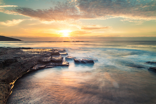 Ocean waves flowing over rocks at sunrise with orange and gold clouds