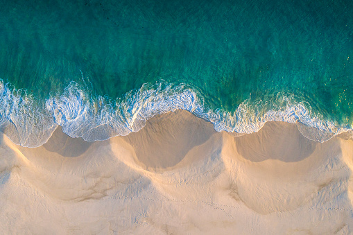 Aerial view of white sand beach coastline and swirling waves with teal blue ocean