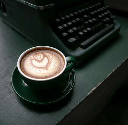 Cup of Flat White coffee in a green cup next to an old typewriter on green wood table. Close up view.