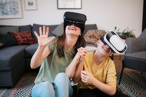 Portrait of a beautiful young Caucasian mother spending some quality time with her young son at home, playing video games using virtual reality googles, having fun and smiling