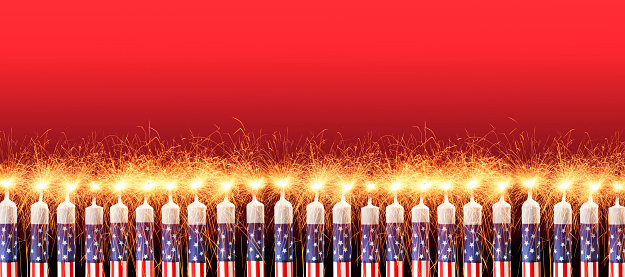A row of ten red, white and blue candles decorated in a patriotic American theme are lit with sparkler flames against a red background.