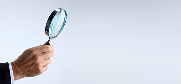 A man wearing a suit holds a magnifying glass in front of a gray background that provides amble room for copy or text.
