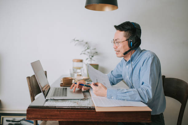 asiatico cinese mid adulto uomo sorridente frequentando lezione online in cucina utilizzando laptop - white collar worker global communications side view headset foto e immagini stock