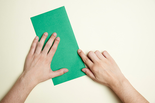 Young hands folding a green square of paper in half to make an origami figure on a light cream colored background. Image with copy space.