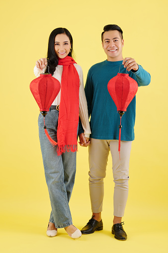 Pretty young Asian boyfriend and girlfriend in warm sweaters holding hands and showing silk lanterns