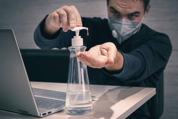 A worker in a medical mask works from home in the early morning. pandemic of covid 19 coronavirus. man treats his hands with a sanitizer to disinfect and destroy dangerous harmful microbes. A young worker in a medical mask works from home in the early morning. pandemic of covid 19 coronavirus. man treats his hands with a sanitizer to disinfect and destroy dangerous harmful microbes. office fogging stock pictures, royalty-free photos & images