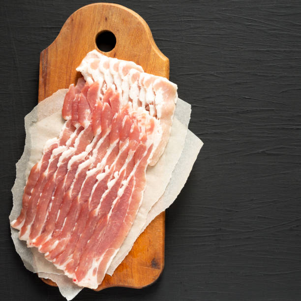 istock Uncooked Raw Bacon on a rustic wooden board on a black background, overhead view. Flat lay, from above, top view. Copy space. 1302397853