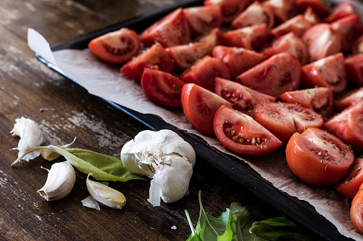 Tomato sliced into halves and fourths, sprinkled with oregano leaves at baking sheet on tray ready for oven slow-roasting drying