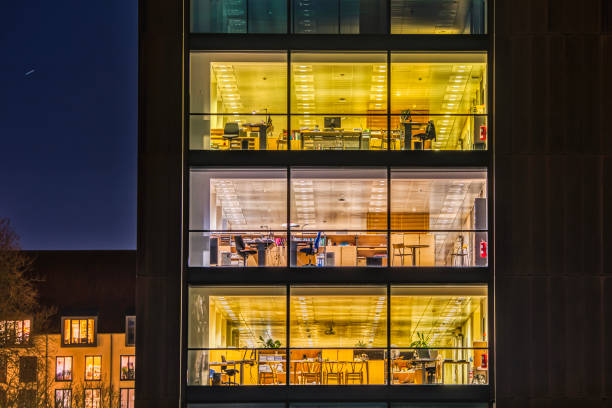 A contemporary office with a broad modern, glazed facade featured by square pattern of wide glazed windows illuminated at night A contemporary office with a broad modern, glazed facade illuminated at night. Square pattern of wide glazed windows of a financial firm exterior. Tech industry headquarter with office space interiors office window night stock pictures, royalty-free photos & images