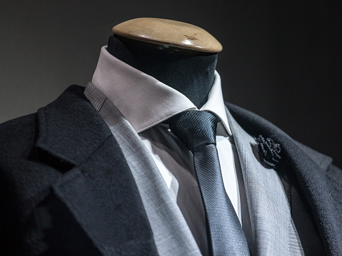 Picture of a formal suit of a man, on a close up, with a grey jacket, a black tie, a black lapel flower pin and a white shirt on display on a dummy in a tailor.