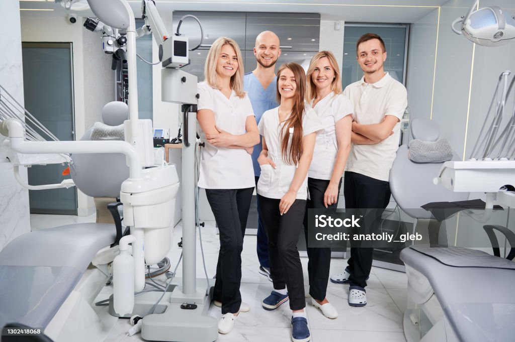 Professional team of dentists and assistants posing in their modern clinic Team of professional dentists and assistants looking at camera and smiling while posing in modern dental office. Dream team. Dentistry and medicine concept. Dental Health Stock Photo