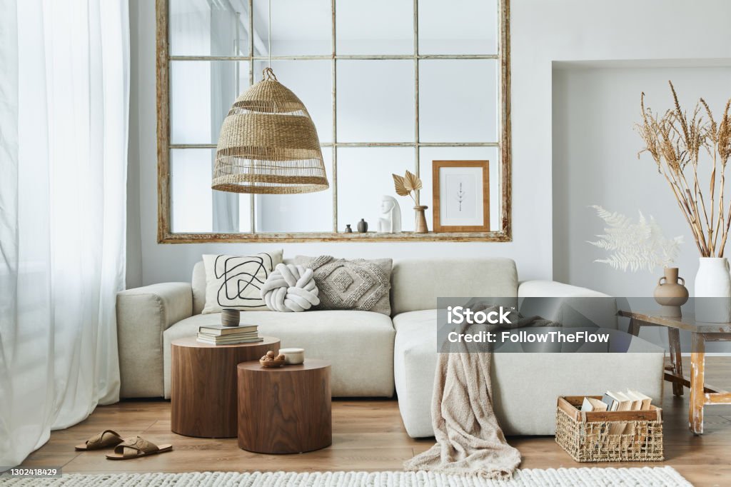 Stylish interior of living room with design modular sofa, furniture, wooden coffee table, rattan decoration, pendant lamp, pillow, dried flowers and elegant accessories in modern home decor. Modern interior of open space with design modular sofa, furniture and elegant personal accessories in stylish home decor. Neutral living room. Living Room Stock Photo