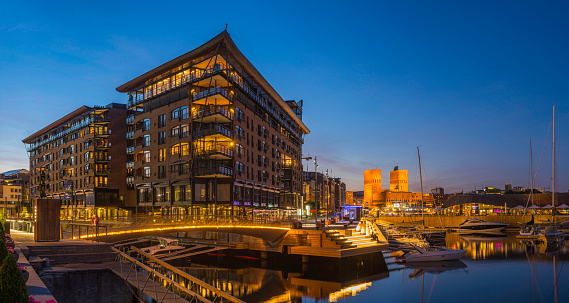 The iconic Functionalist towers of Oslo City Hall, Radhus, spotlit against the chrome blue dusk sky overlooking the crowded promenades and marinas of Aker Brygge, the popular leisure, dining and exclusive residential district in the heart of downtown Oslo, Norway's picturesque capital city.