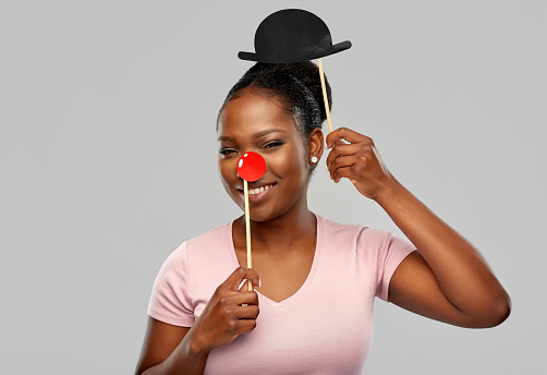 red nose day, party props and photo booth concept concept - happy african american young woman with clown nose over grey background