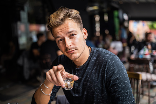 Portrait of a young man looking at the camera. He is sitting in a coffee bar and he is holding a cup of coffee showing with his finger at the camera.
