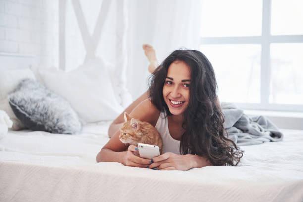 istock Using smartphone. Attractive blonde resting on the white bed with her cute kitten 1302423009