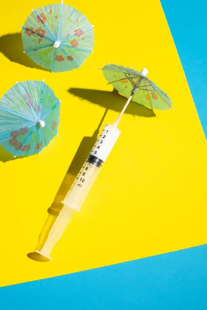 Syringe with cocktail umbrella on the beach made of yellow and blue paper. Summer 2021 tourism and traveling concept. Minimal Coronavirus or 2019-nCoV or COVID-19 scene.