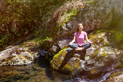 Woman sitting on a rock in the middle of nature, being sunbathed doing yoga and meditation