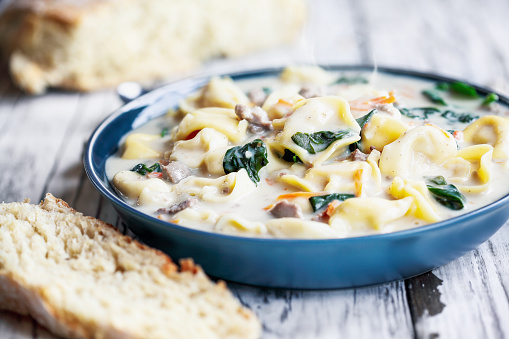 Fresh Tortellini Soup with Italian sausage, spinach and carrots. Served with homemade artisan bread over a white wood wooden table. Selective focus with blurred background.