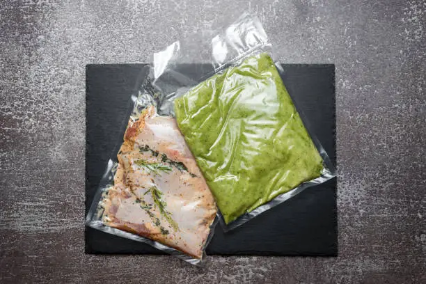 Products in vacuum packaging on black slate board. Chicken meat with herbs and green beans puree, vacuum sealed food ready for sous vide cooking. Sous-vide, new technology cuisine in quarantine time.