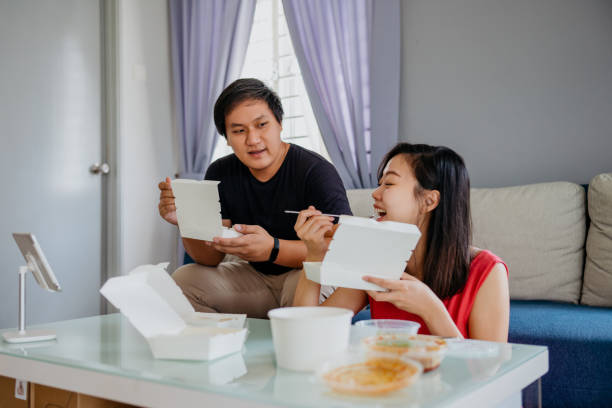 Asian Chinese couple having take out food at home together Image of an Asian man and woman having take out food at home together and using digital tablet for video calling or watching a show. asian food delivery stock pictures, royalty-free photos & images