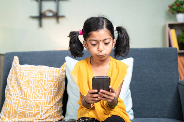 istock Young girl kid using mobile phone while sitting on sofa in bending head posture - concept of Kid mobile phone game addiction, technology and modern lifestyle. 1302433910