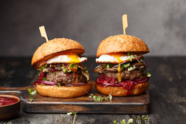 Junk food - homemade beef burgers on vintage wooden background Two homemade beef burgers with mushrooms, micro greens, red onion, fried eggs and beet sauce on wooden cutting board. Side view, close up fine dining stock pictures, royalty-free photos & images