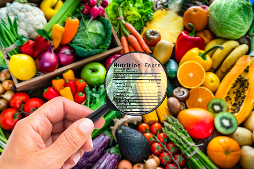 Close up view of a female hand holding magnifying glass on a colorful group of healthy fruits and vegetables background to look for the details of the nutrition facts of this type of food. A Nutrition Facts label is visible on the magnifying glass.