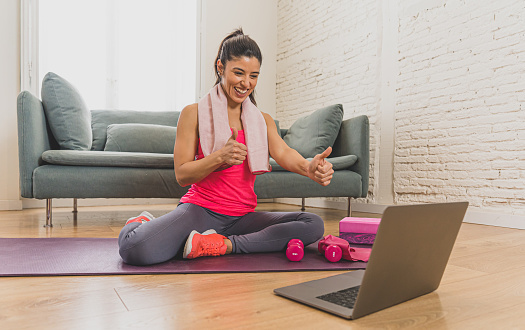 Young Beautiful latin Woman working out at home connected to online fitness class on the computer laptop. Woman personal trainer coach on zoom teaching exercises in live streaming workout class.